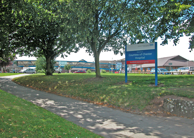 The entrance to the Countess of Chester Hospital seen from a footpath across a grass verge, with a blue NHS sign reading “Countess of Chester Hospital — Accident and Emergency” beside mature trees and low hospital buildings.
