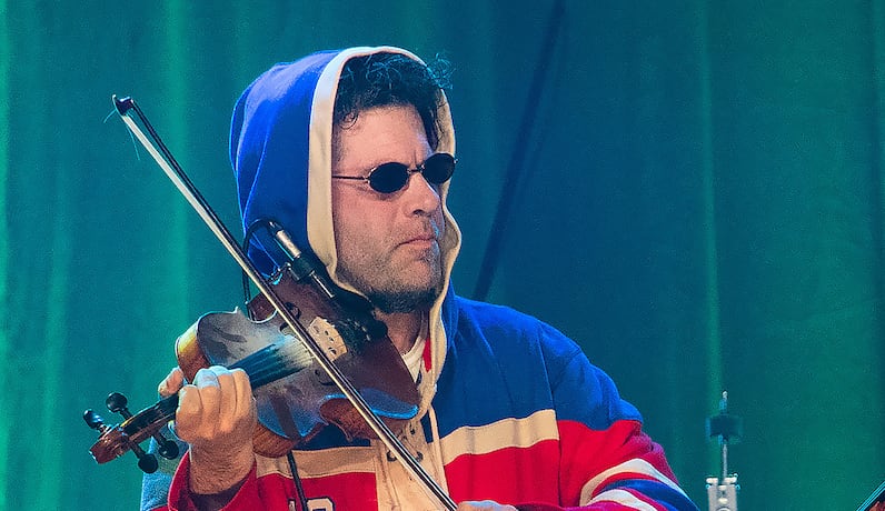 Two people play fiddles onstage in a performance. The man on the left is wearing a hockey jersey and sunglasses, while the man on the right is wearing a suit.