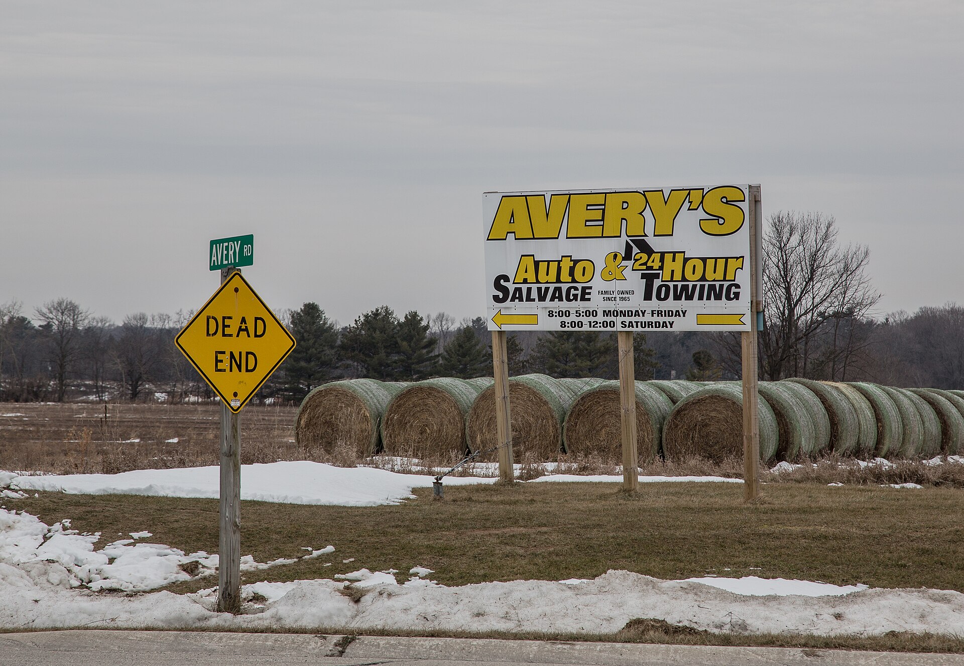 A roadside sign for Avery's Auto Salvage & 24 Hour Towing stands in a snowy field lined with hay bales, beside a green Avery Rd street sign and a yellow Dead End sign, in Two Rivers, Wisconsin.