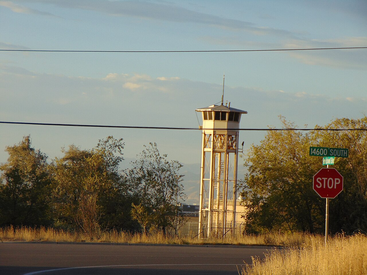 A tall white guard tower on steel legs rises above trees and dry grass beside a road junction with a stop sign and a 14600 South street sign, at the former Utah State Prison in Draper.