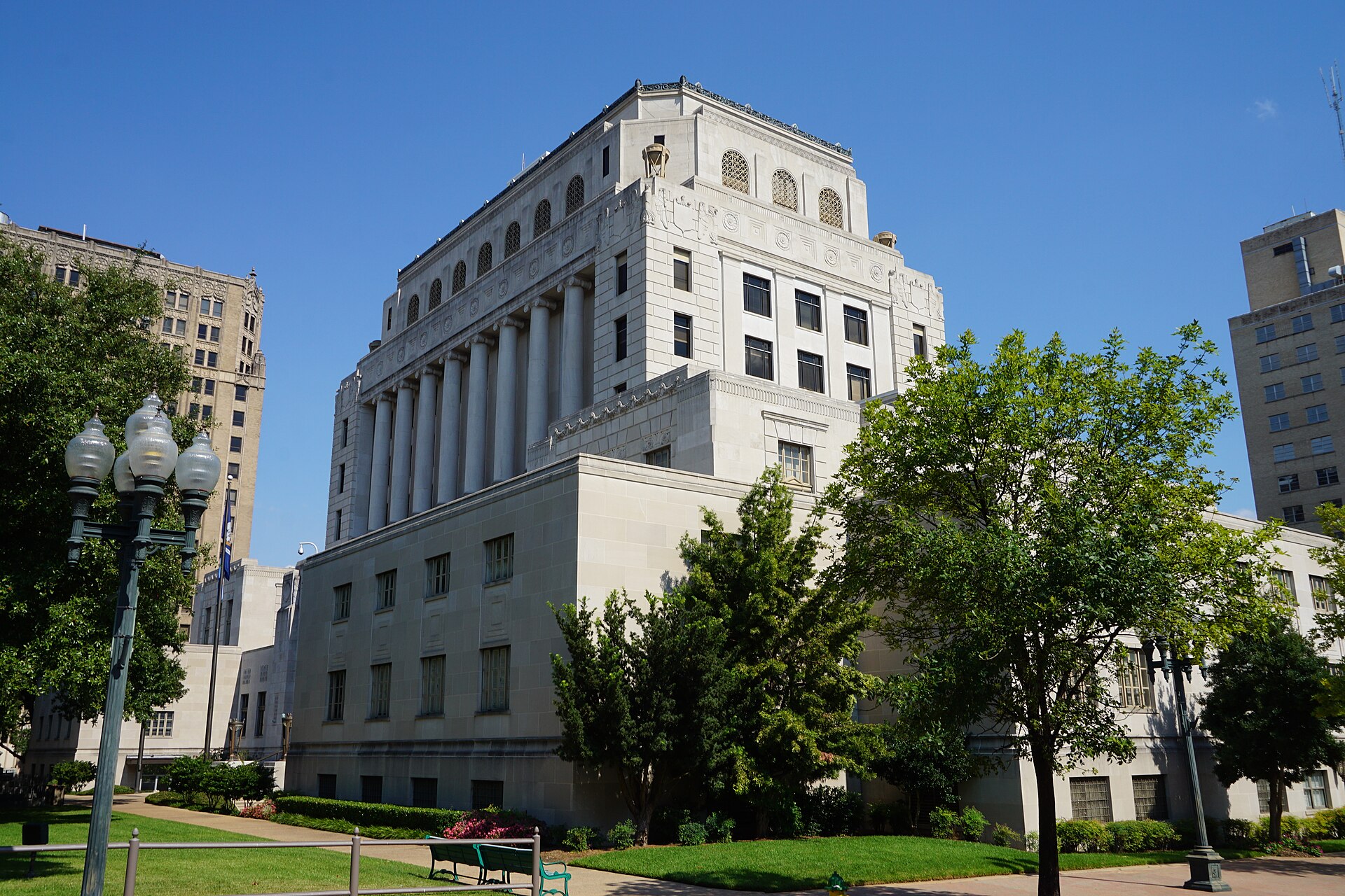 The pale stone Caddo Parish Courthouse in Shreveport, Louisiana, a tall stepped building with a row of columns, seen across lawns and young trees under a deep blue sky.