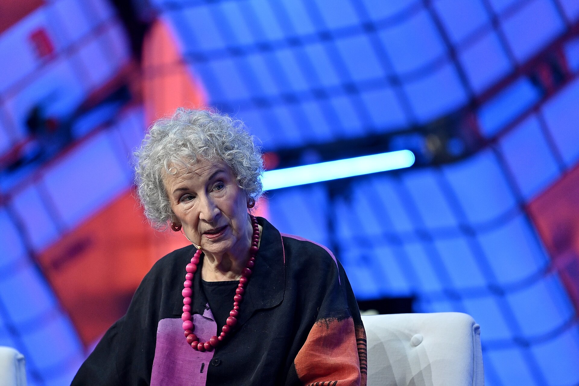 Margaret Atwood, with curly grey hair and a long red bead necklace, seated on stage speaking, against a blurred red and blue lit backdrop.