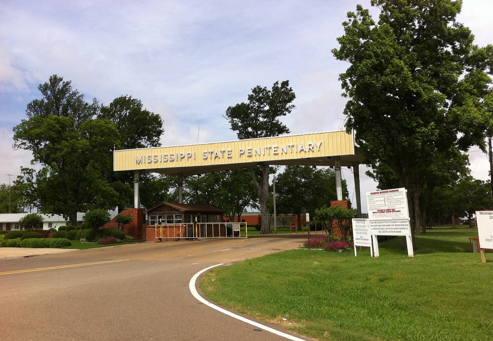 The main entrance of the Mississippi State Penitentiary at Parchman: a pale yellow gantry reading MISSISSIPPI STATE PENITENTIARY spans the road above a brick guardhouse, with tall trees, lawns and warning signs on either side.