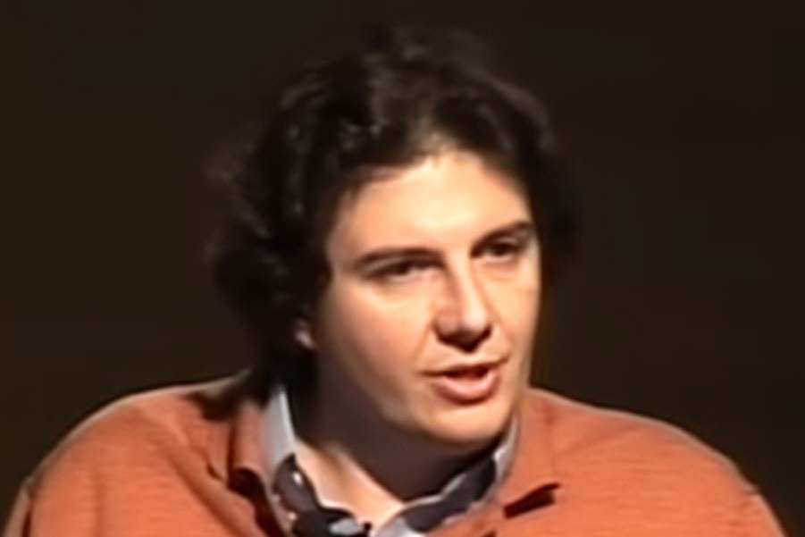 David Sabatini, with dark curly hair, in an orange sweater over a collared shirt, speaking at a lecture against a dark background.