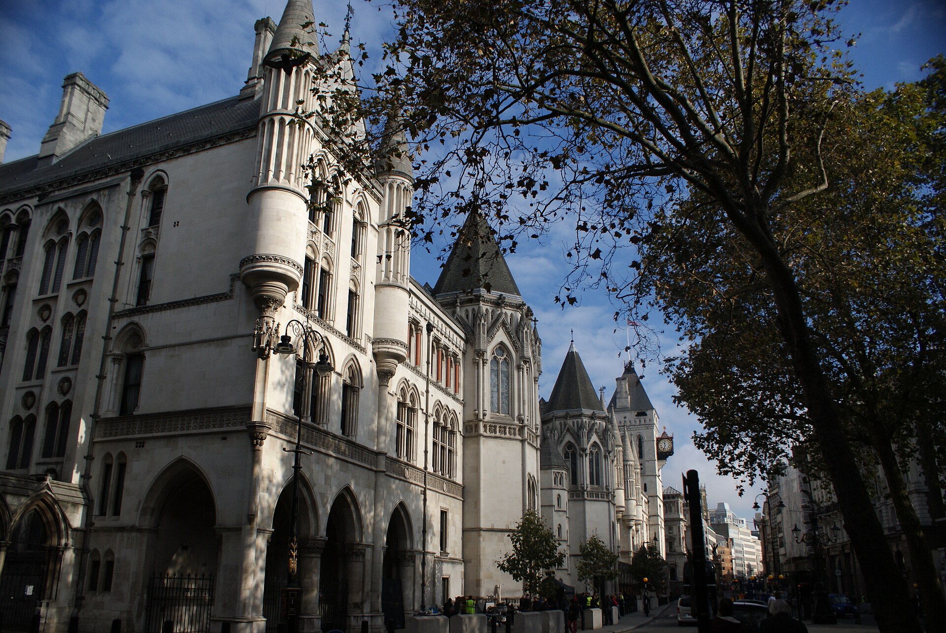 The pale stone Gothic Revival frontage of the Royal Courts of Justice on the Strand, London, with turrets and a clock tower receding down the street under a blue sky, framed by autumn plane trees.