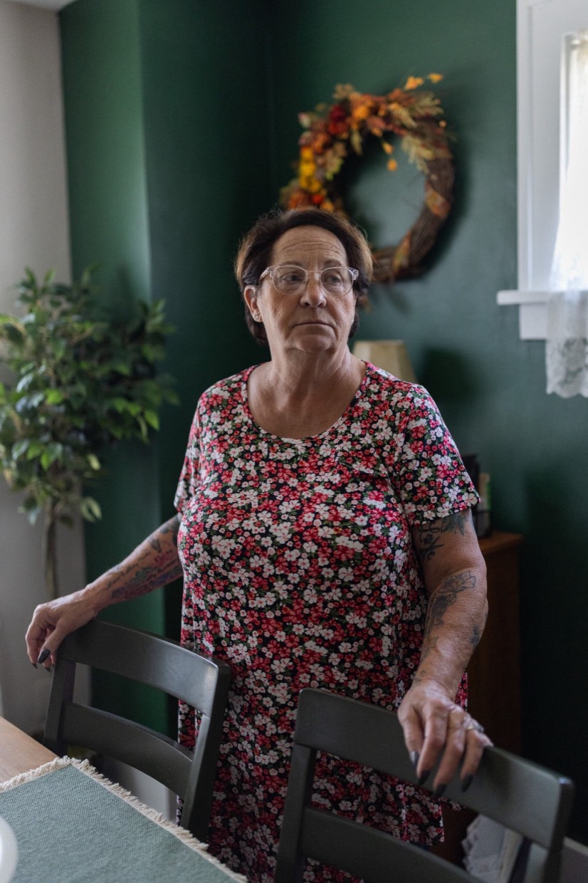 Nancy Smith, wearing glasses and a floral-pattern T-shirt stands by a dining table in front of a green wall.