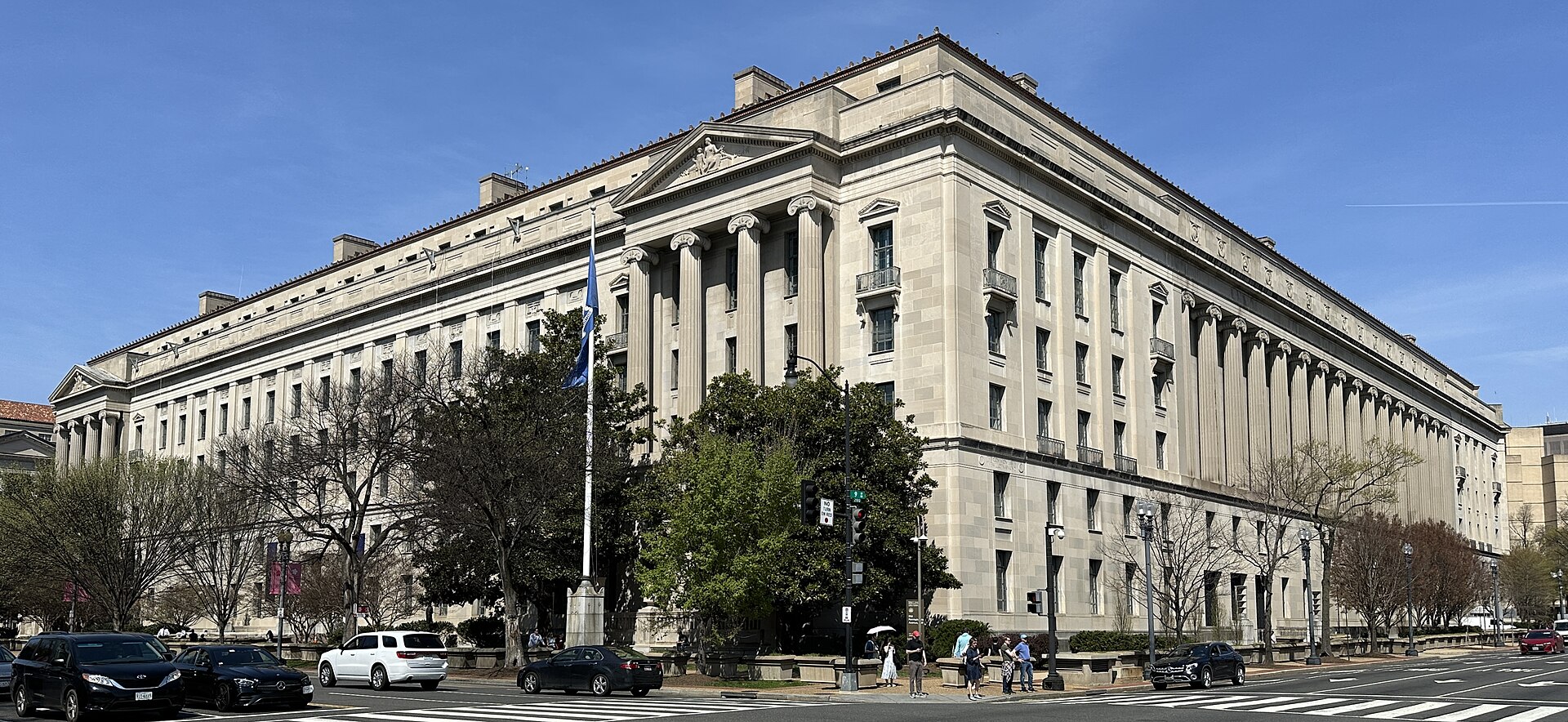 The Robert F. Kennedy Department of Justice Building in Washington, D.C., a monumental limestone building with Ionic columns and a pedimented entrance, photographed from a street corner under a clear blue sky.