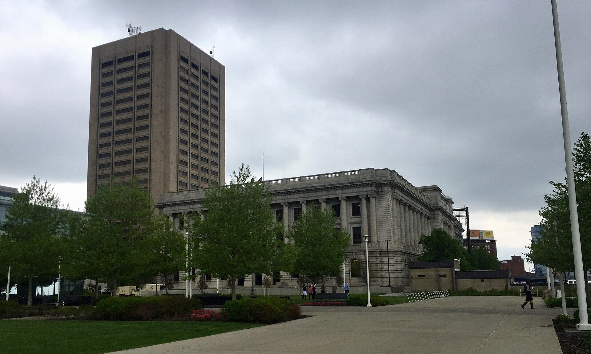 The grey Beaux Arts Cuyahoga County Courthouse in Cleveland behind a row of young trees, with a plain concrete office tower rising behind it under a heavy overcast sky.