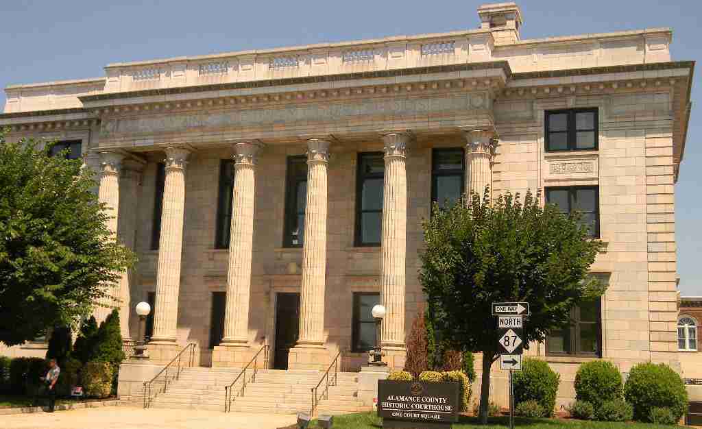 The Alamance County Historic Courthouse, a pale stone building with a row of fluted columns above wide front steps, seen on a sunny day behind trees and a sign reading One Court Square.