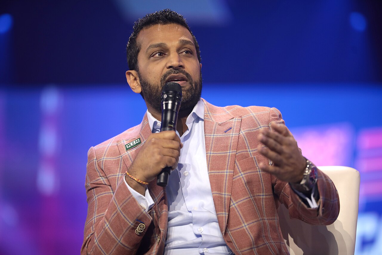 Kash Patel, seated and gesturing as he speaks into a handheld microphone, wearing a salmon checked jacket over an open-collared white shirt against a blue stage backdrop.