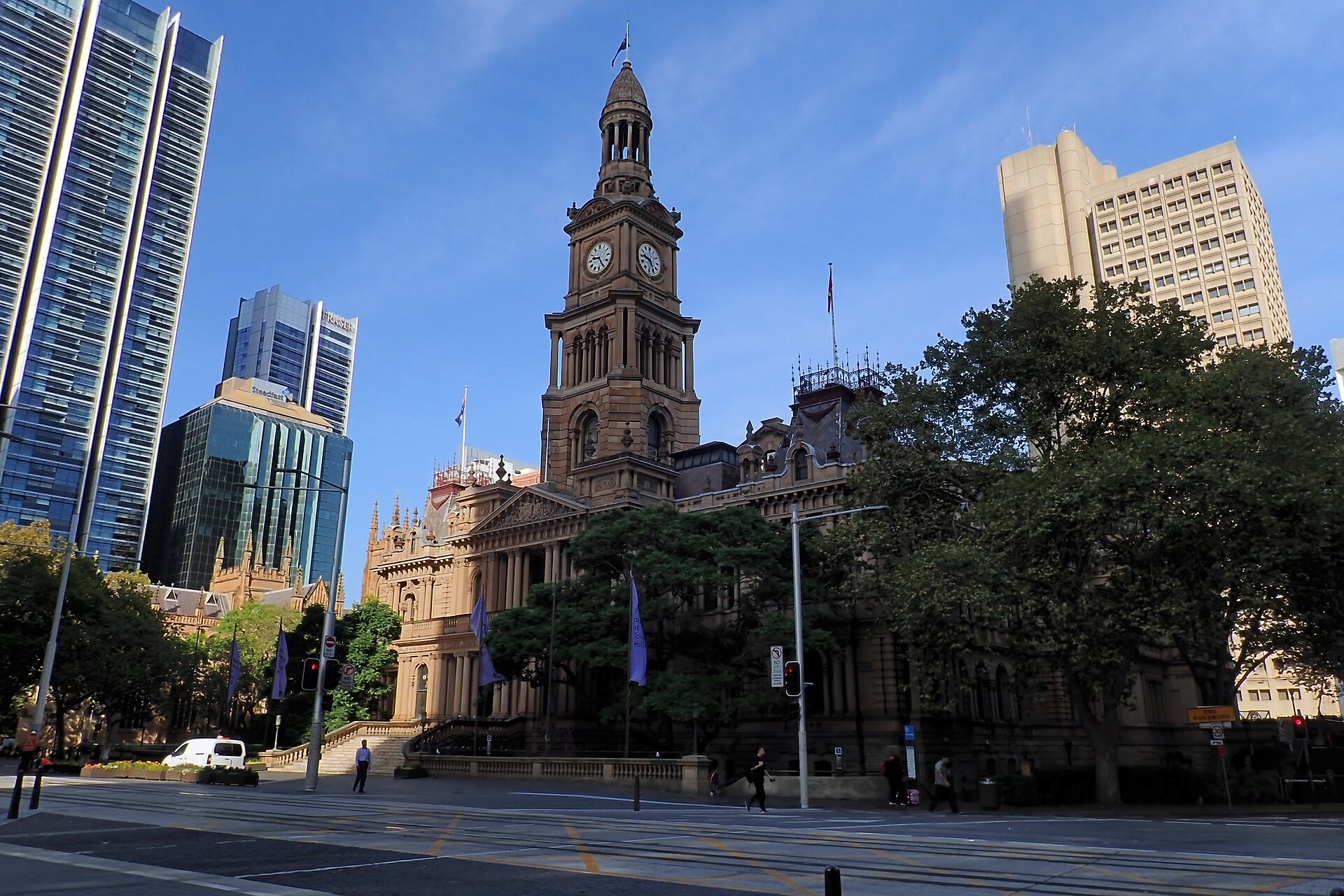 Sydney Town Hall on George Street, a sandstone Victorian building with a tall clock tower, flanked by trees and modern office towers under a clear blue sky.