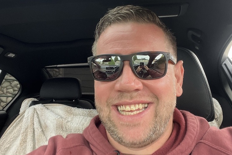 Jordan Coleman, smiling in sunglasses and a maroon hoodie, taking a selfie in the driver's seat of a car.