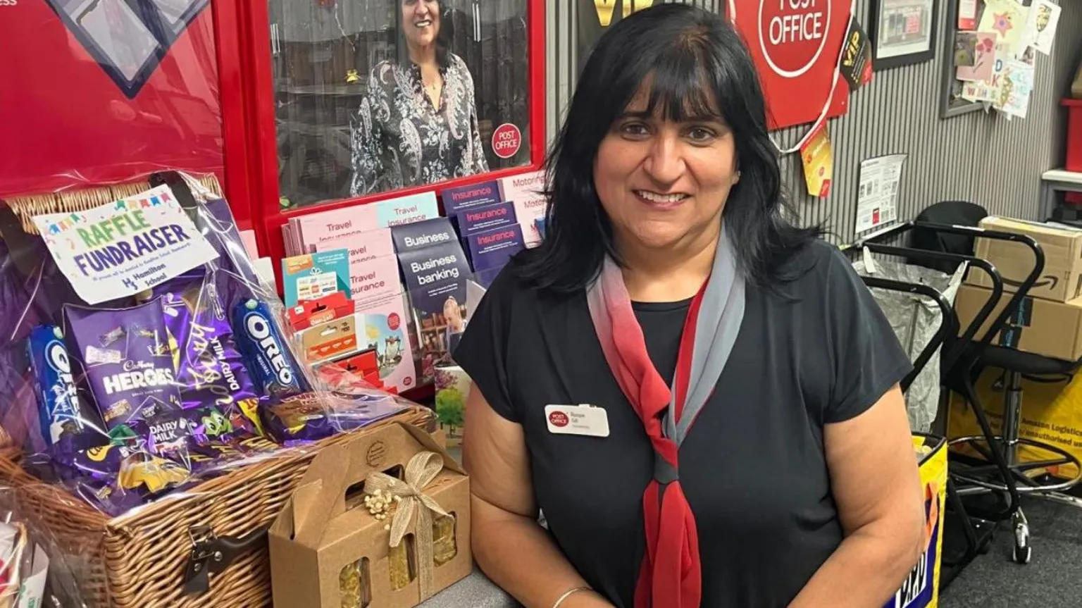 Rooprit Gill standing behind a Post Office counter beside a raffle fundraiser display of a wicker hamper, chocolates and biscuits, with Post Office signage and community noticeboards in the shop behind her.
