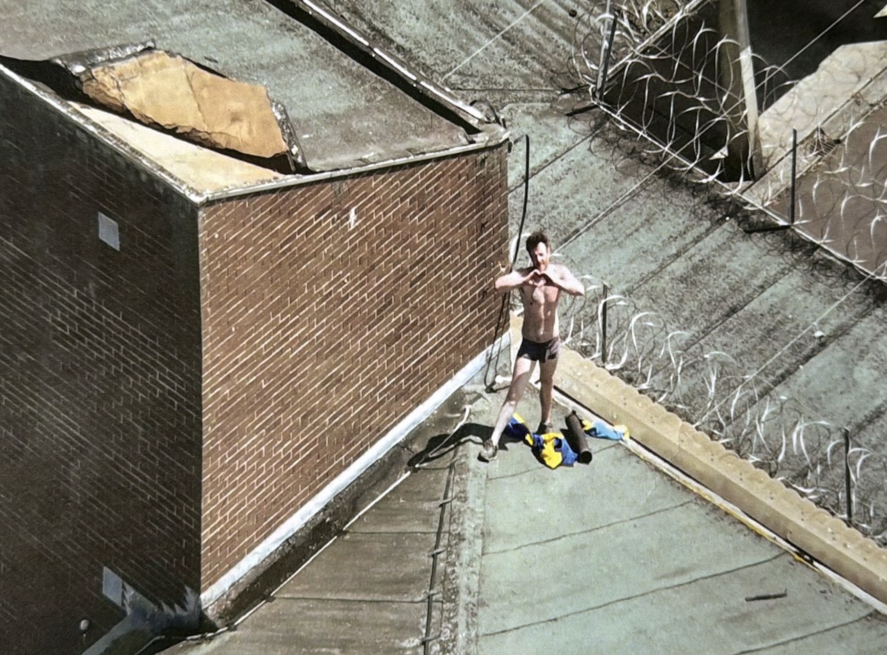 Aerial photograph of Joe Outlaw, shirtless and in his underpants, standing on a prison roof beside coils of razor wire and making a heart shape with his hands.