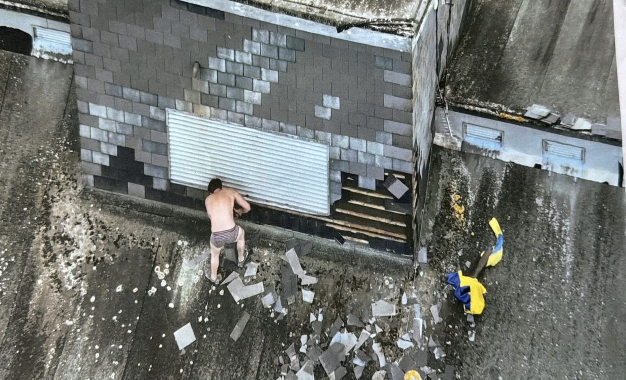 Aerial photograph of Joe Outlaw in his underpants on the prison roof, pulling at a metal shutter amid scattered broken roof tiles.
