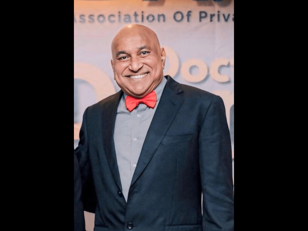 Dr James Jeremiah smiling in a dark suit, grey shirt and red bow tie, standing in front of an Association of Private Practitioners Sabah banner.