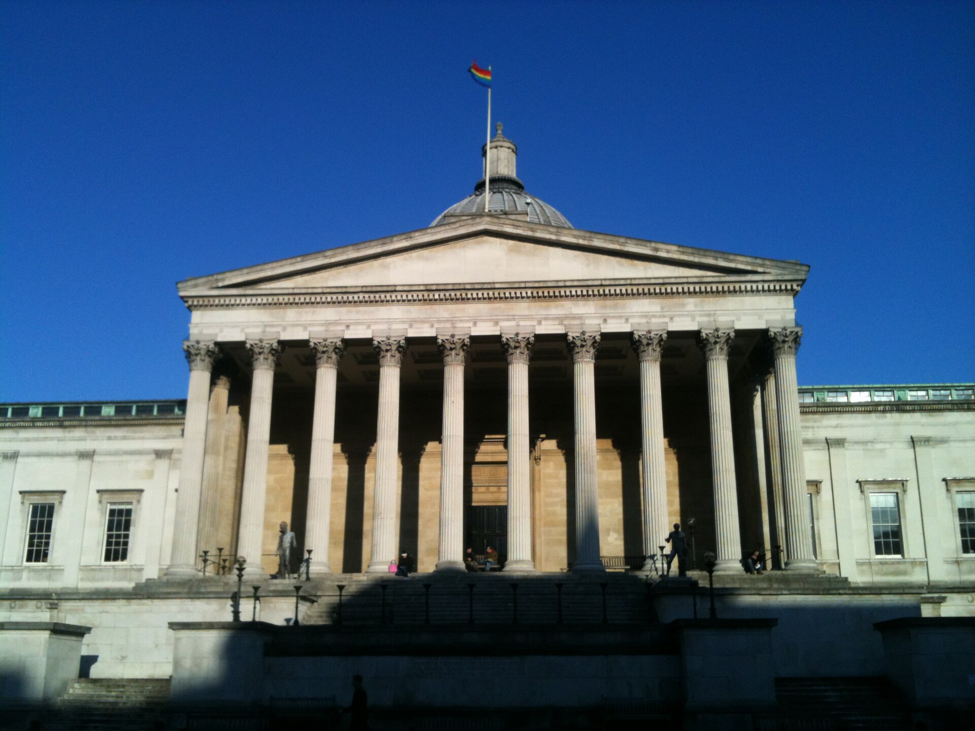 The neoclassical Wilkins Building at University College London, seen head-on from the quad - a wide Corinthian portico of ten fluted columns beneath a pediment, with a domed roof behind it against a deep blue sky.