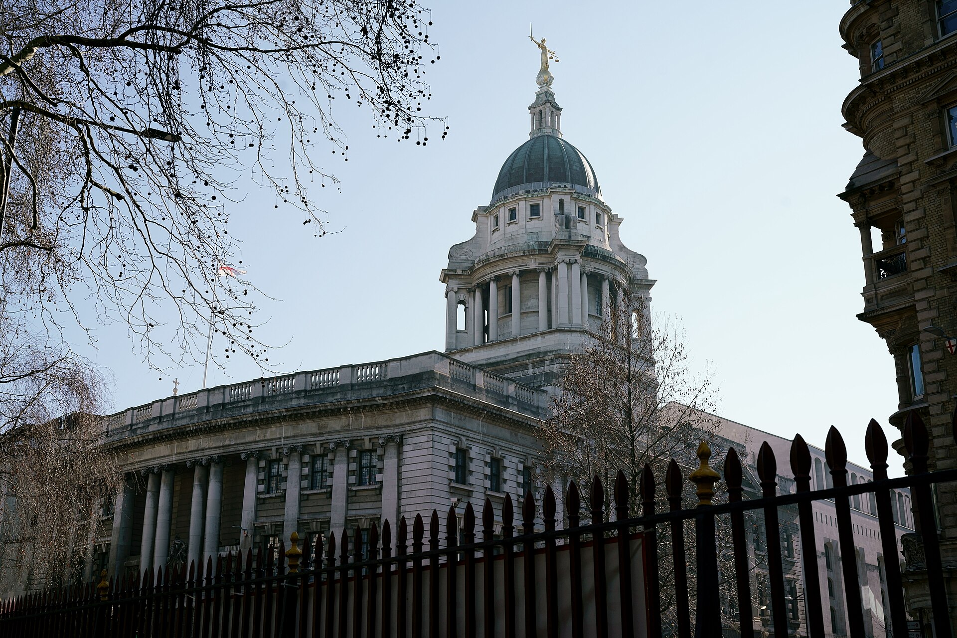 The Central Criminal Court in London seen from behind railings on a winter afternoon, its stone dome topped by the gilded figure of Lady Justice holding scales and a sword, with bare tree branches in the foreground.