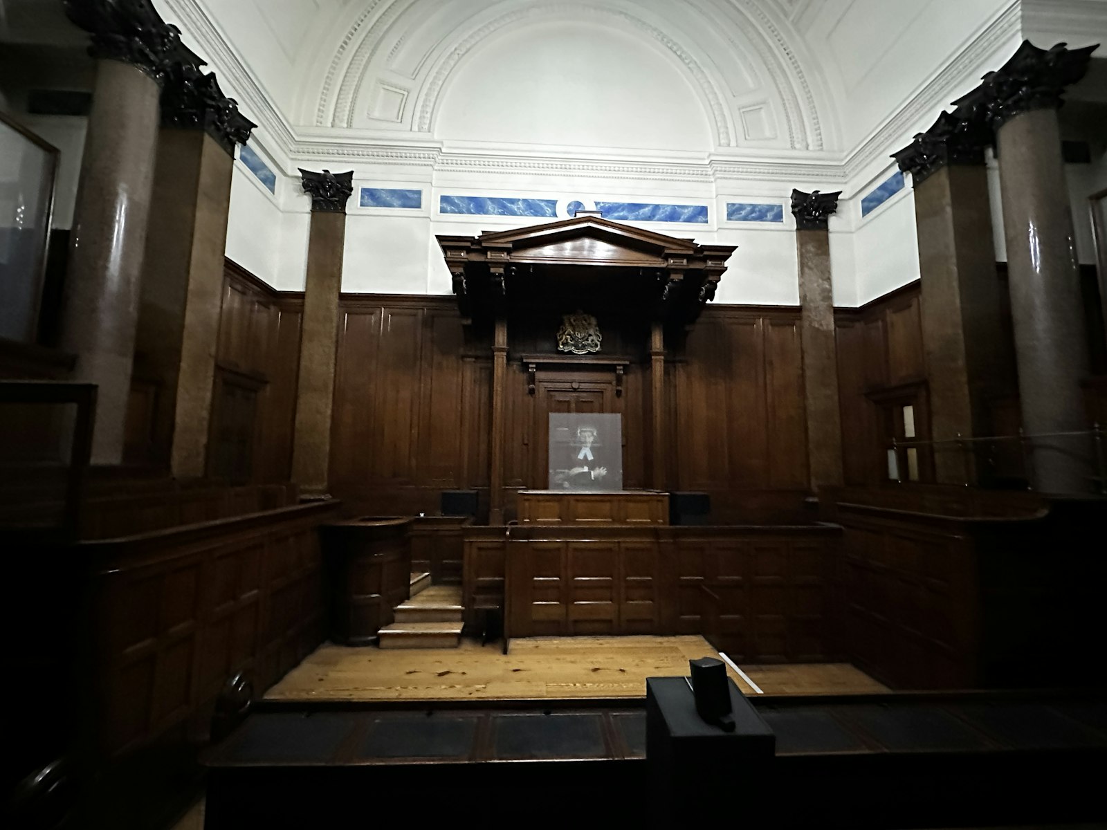 An empty, dimly lit courtroom with dark wood panelling, marble columns and a canopied judge's bench beneath a royal coat of arms.