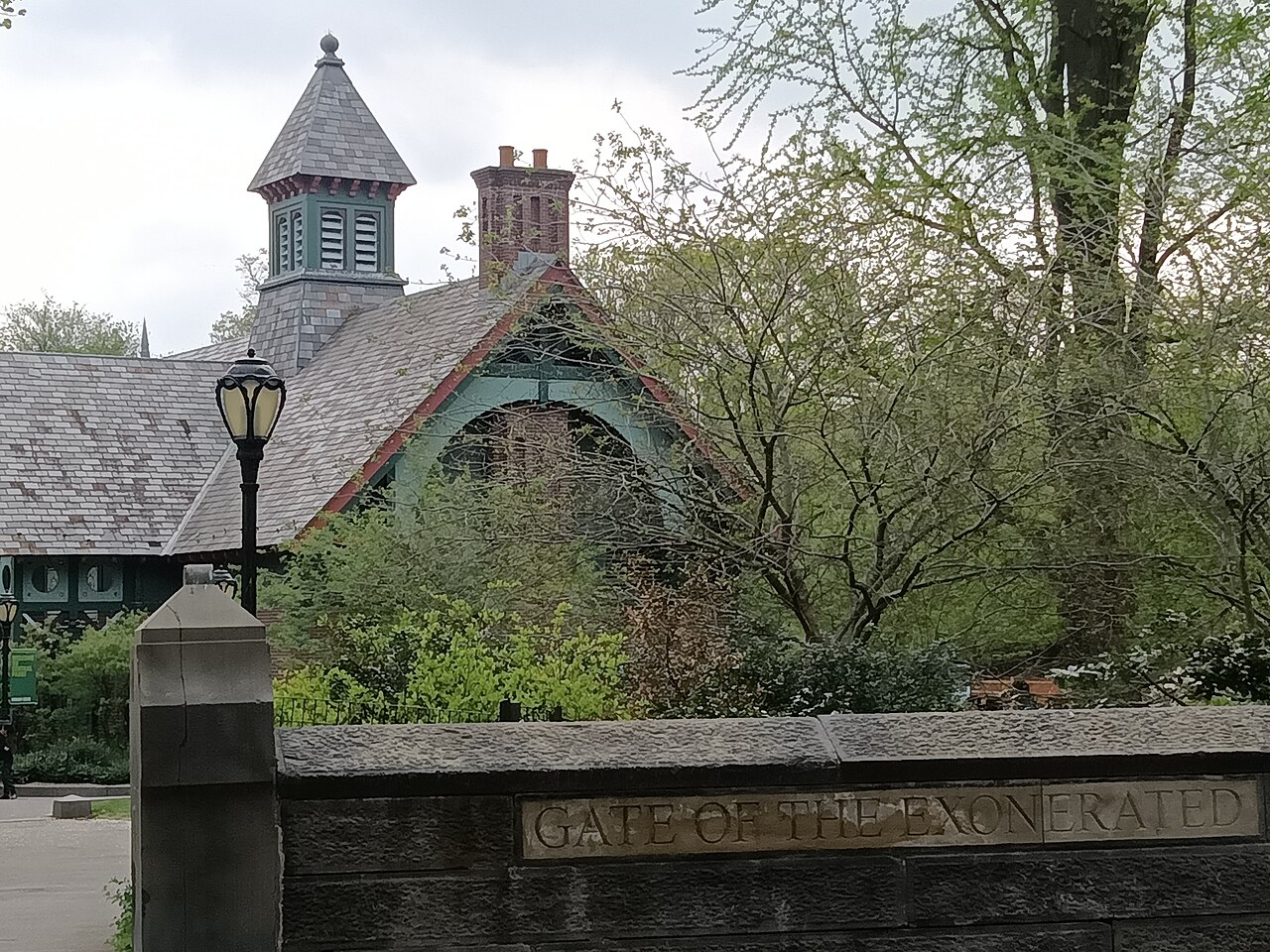 A carved stone inscription reading GATE OF THE EXONERATED along a low wall at an entrance to Central Park, with a gabled park building and bare spring trees behind it.