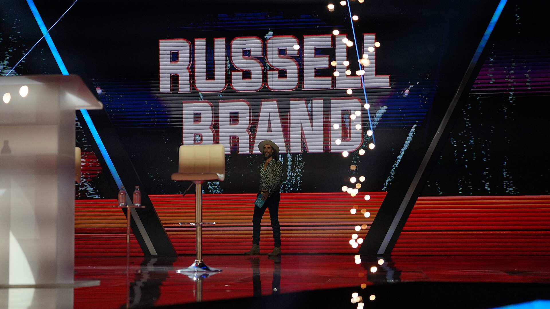 Russell Brand, in a hat and patterned shirt, stands on a red-lit stage beneath a giant screen spelling out his name, with an empty bar stool and a lectern in the foreground.