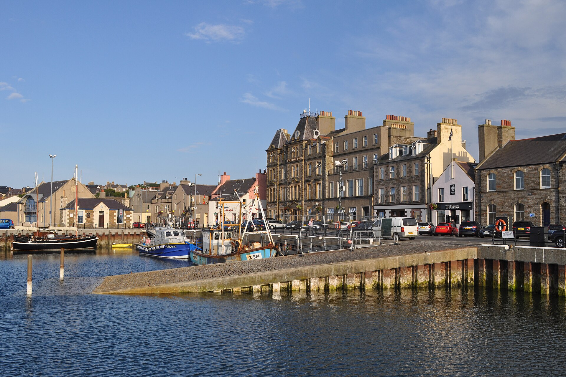 Kirkwall harbour in Orkney on a sunny evening, fishing boats moored beside a cobbled slipway in front of the stone-built Kirkwall Hotel and harbour-front houses.