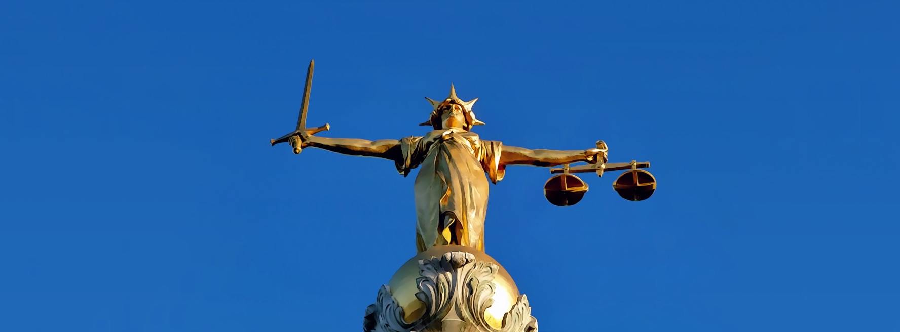 The gilded Lady Justice statue atop the Old Bailey in London, holding a raised sword in one hand and a set of scales in the other, against a clear blue sky.