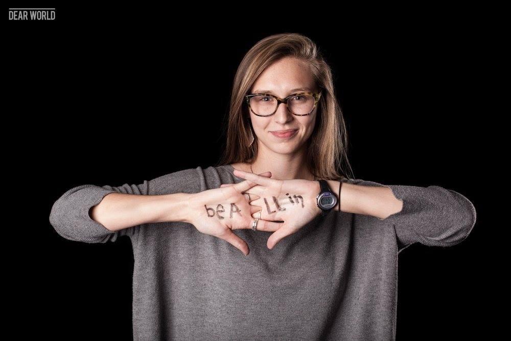 Madison Reddick in a grey sweater against a black background, with writing on her hands, in a Dear World portrait