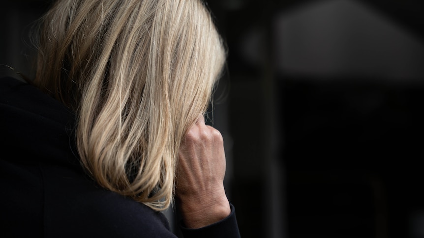 A woman with shoulder-length blonde hair, seen from behind, holding a phone to her ear.