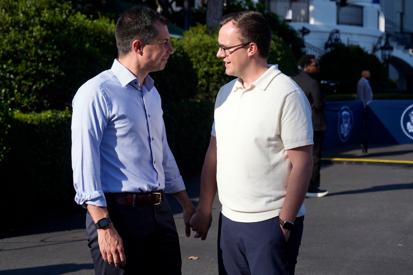 Pete Buttigieg, in a light blue shirt with rolled sleeves, and his husband Chasten, in a cream polo shirt and glasses, stand facing each other on the White House grounds.