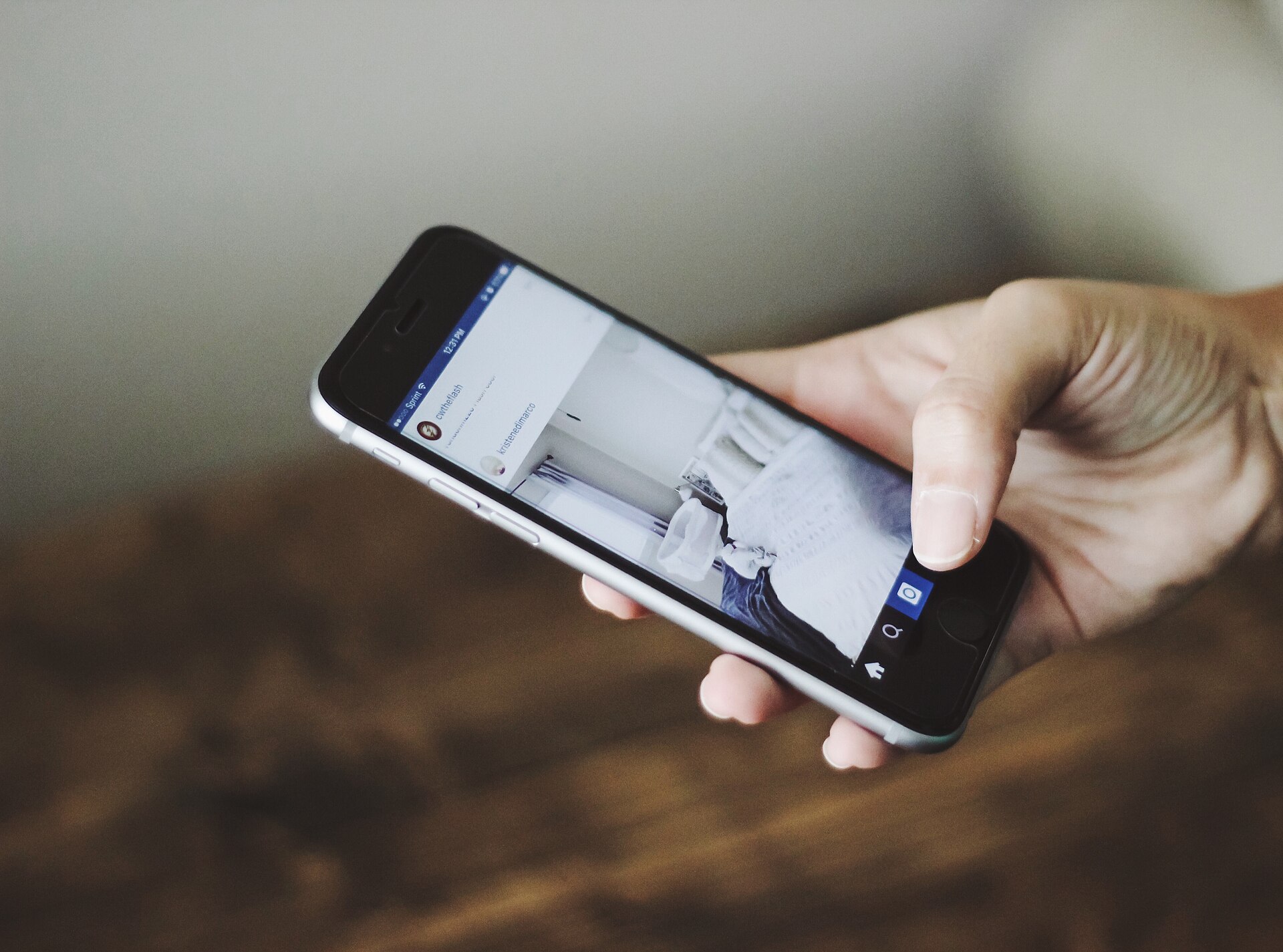 A hand holding a smartphone open on the Instagram app, against a blurred grey and brown background.