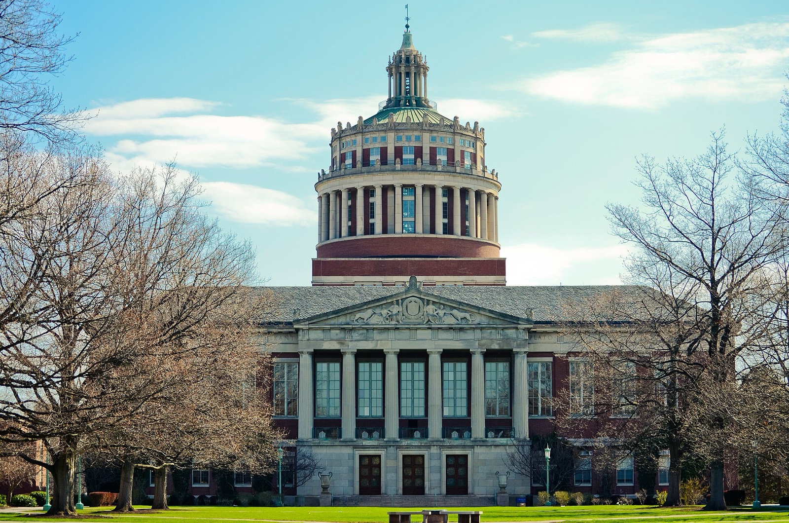 Rush Rhees Library at the University of Rochester: a pale stone portico with tall columns and a pediment, beneath a red-brick and stone rotunda topped by a green copper cupola, framed by bare trees under a blue sky.