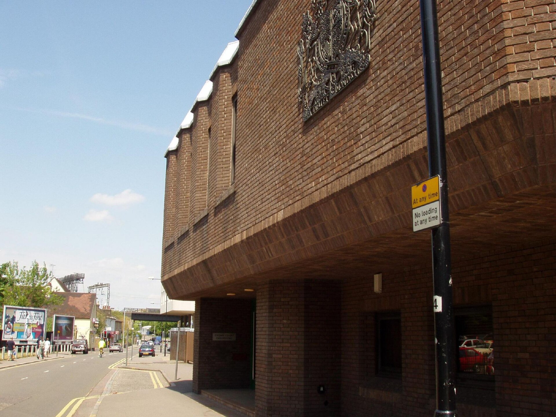 The brown-brick frontage of Chelmsford Crown Court, its upper storey overhanging the entrance and bearing a large metal royal coat of arms, beside a street running down to a railway bridge.