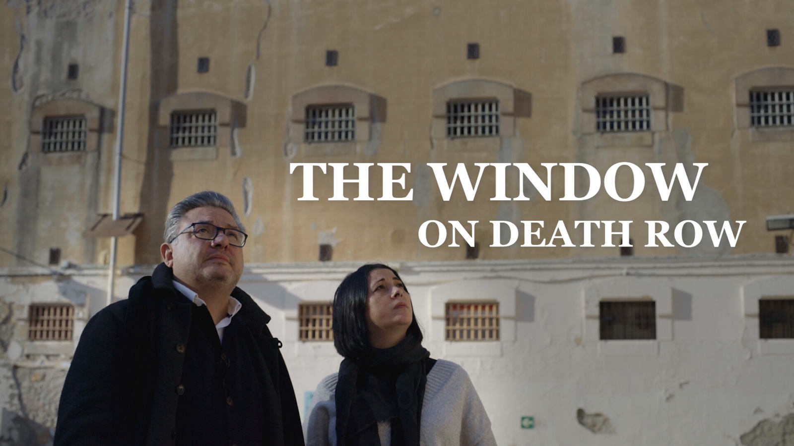 Title card for The Window on Death Row: Joaquin Jose Martinez, a man with grey hair and glasses in a dark coat, and his wife Jessica, in a dark scarf, look up at the barred windows of the old La Modelo prison in Barcelona.