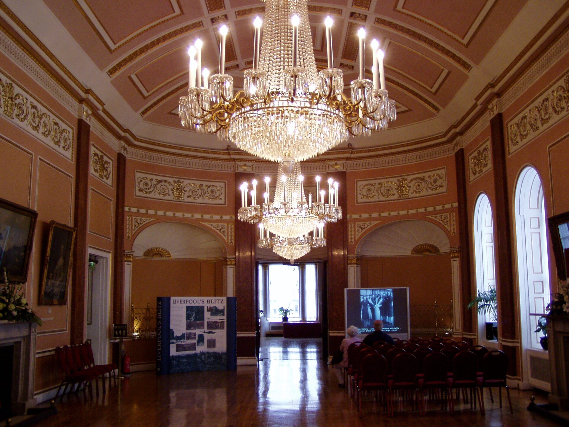 A chandeliered reception room in Liverpool Town Hall, its coffered ceiling hung with two large crystal chandeliers above a polished wooden floor, with rows of red chairs set out at one end beside free-standing display boards.