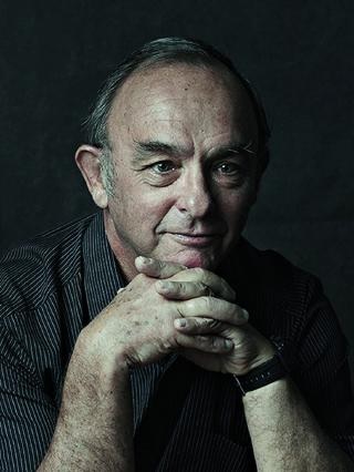 Studio portrait of Claude Imhoff, grey-haired and in a pinstriped jacket, resting his chin on clasped hands against a dark background.