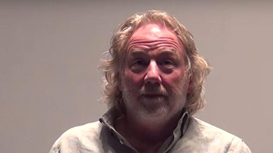 Timothy Busfield, with long curly grey-blond hair and a beard, wearing a pale linen shirt, speaking against a plain grey background.