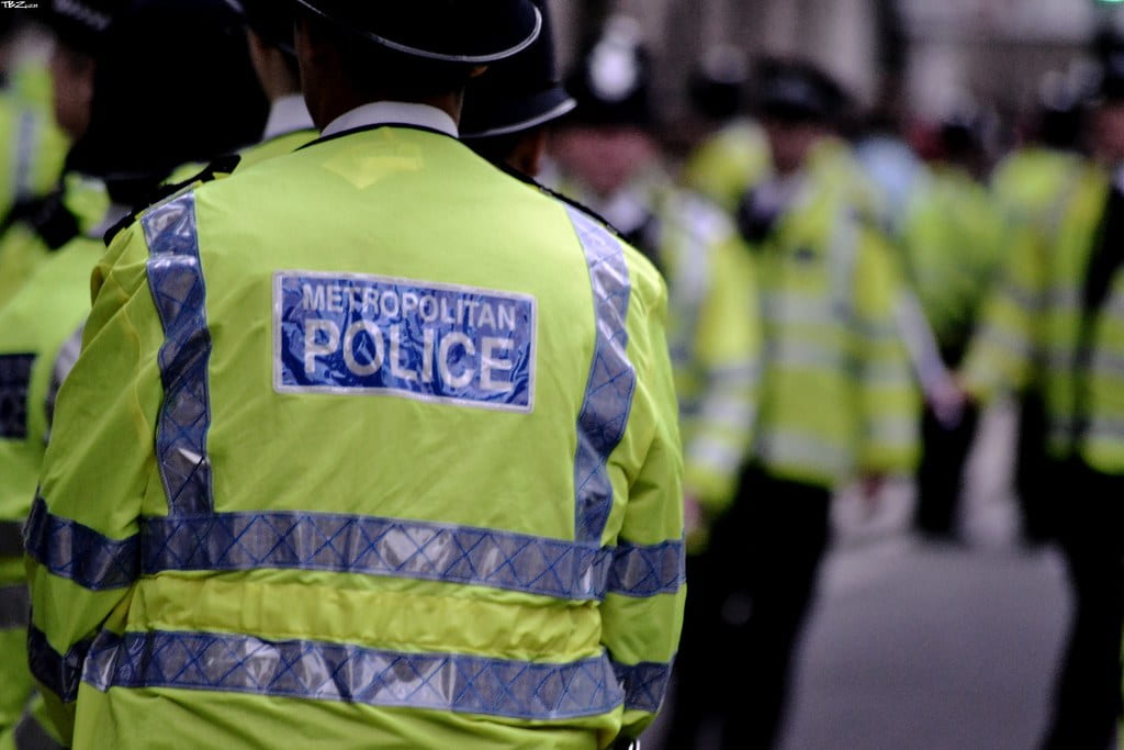A line of Metropolitan Police officers in hi-vis jackets and custodian helmets, photographed from behind on a London street.