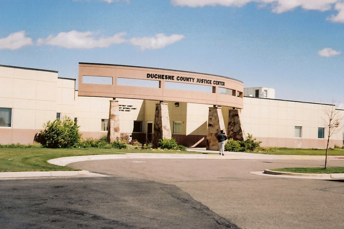 The low cream-coloured Duchesne County Justice Center in Duchesne, Utah, with a pink concrete arch on stone pillars bearing its name over the entrance, a lawn and a curved driveway.
