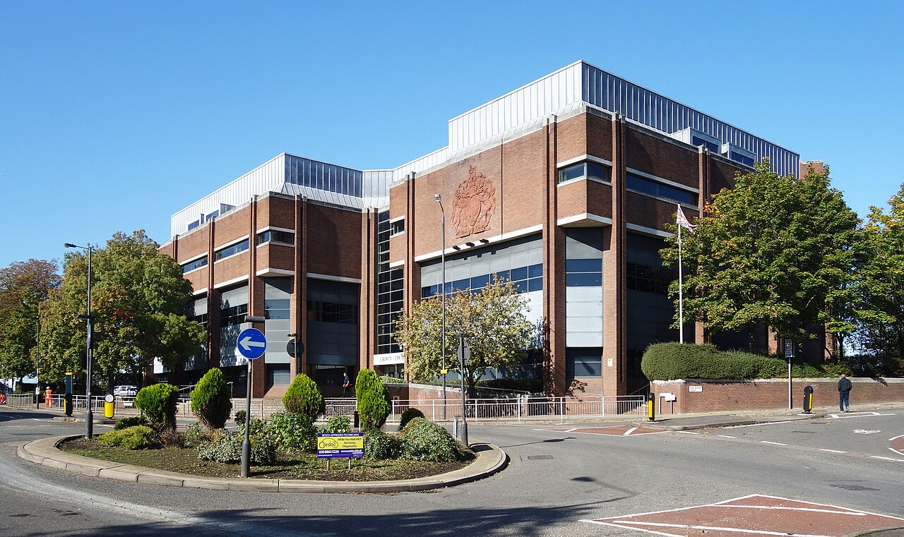 Harrow Crown Court, a red-brick and glass court building of the 1970s with a stone Royal coat of arms in relief on its upper facade, seen from a road junction under a clear blue sky.