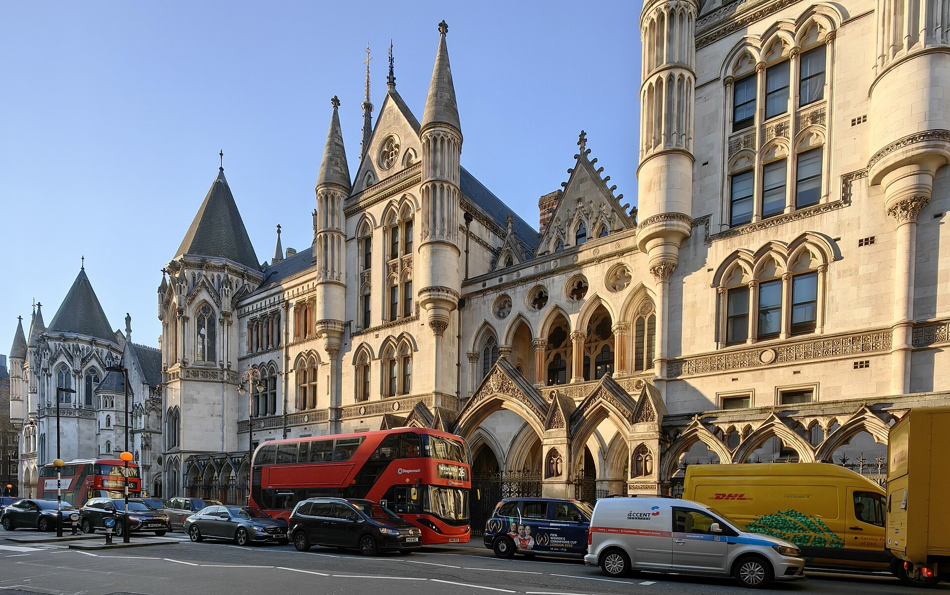 The Royal Courts of Justice on the Strand in London, a Victorian Gothic stone building with pointed arches, turrets and spires, with a red double-decker bus and traffic passing in front under a clear blue sky.