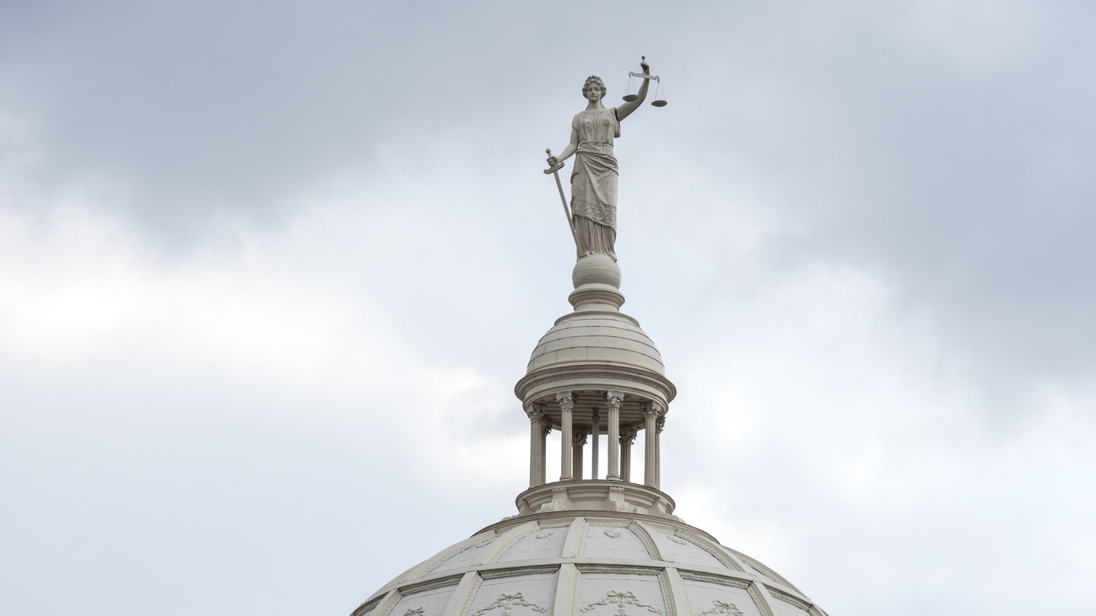 A white statue of Lady Justice holding a sword and raised scales atop the columned lantern and dome of the McLennan County Courthouse in Waco, Texas, against a grey cloudy sky.