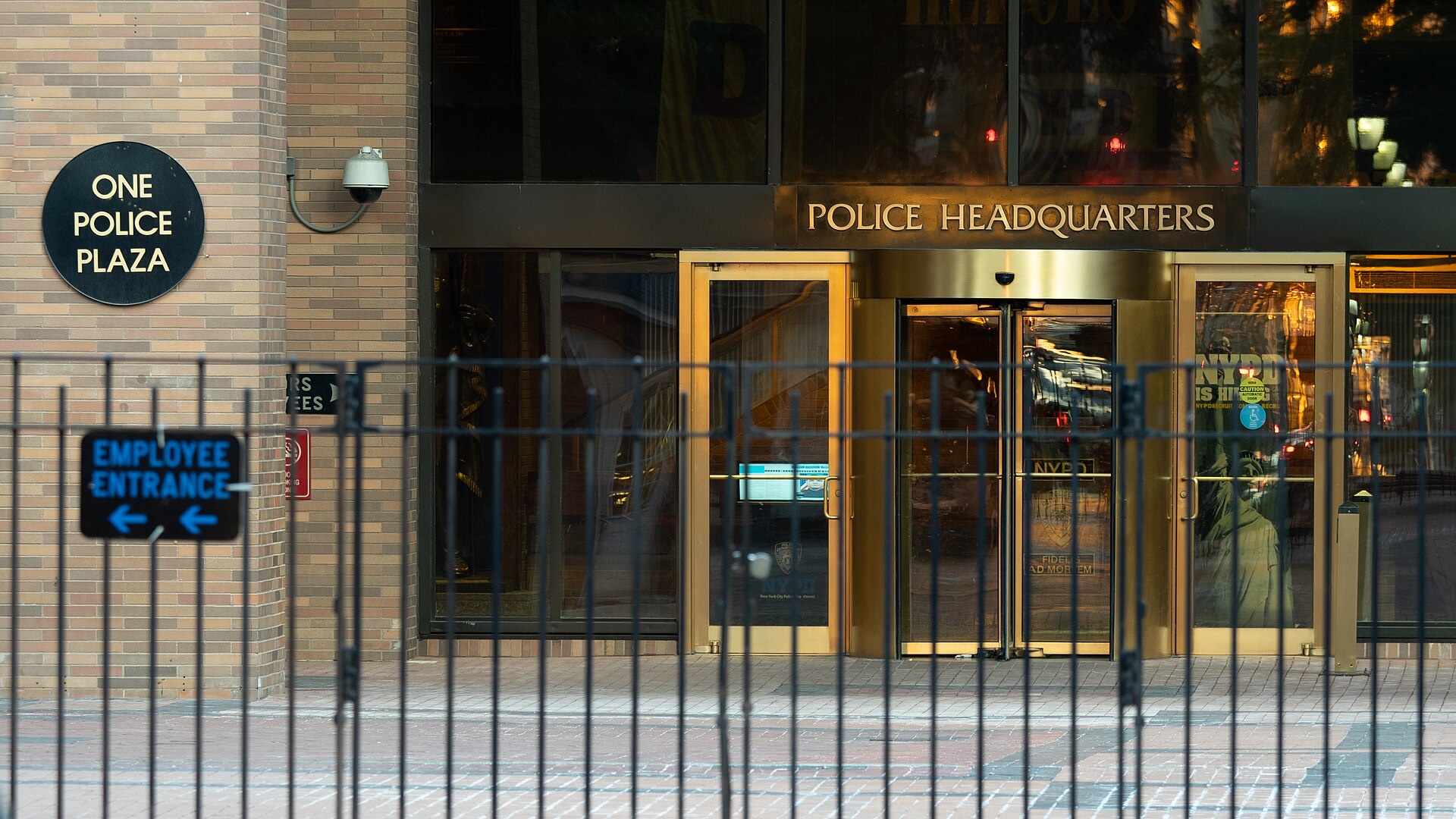 The entrance of NYPD headquarters behind a black iron fence, with a round 'One Police Plaza' plaque on the brick wall, a blue 'Employee Entrance' sign and 'Police Headquarters' lettered above glass doors.